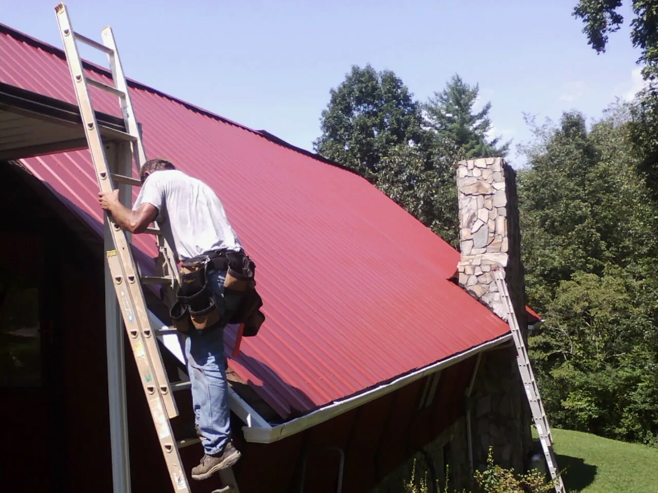 Roofer on a ladder beside a red metal roof and stone chimney — Soffit & Fascia Repair by Roofing Brothers in Stanwood